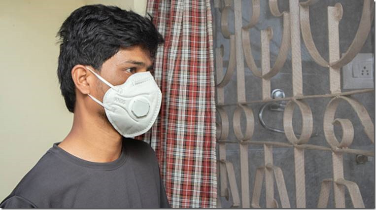 Young man with medical face mask watching through window - concept of Home isolation or quarantine due to covid 19 or coronavirus outbreak